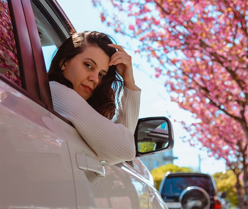 A brown hair girl looking through the window of a white car in front of a blooming tree with pink flowers.