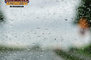 Raindrops covering a car windshield with blurred road and scenery, featuring the Stewart’s Automotive on Donnybrook logo in the corner.