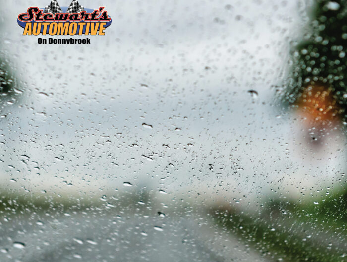 Raindrops covering a car windshield with blurred road and scenery, featuring the Stewart’s Automotive on Donnybrook logo in the corner.