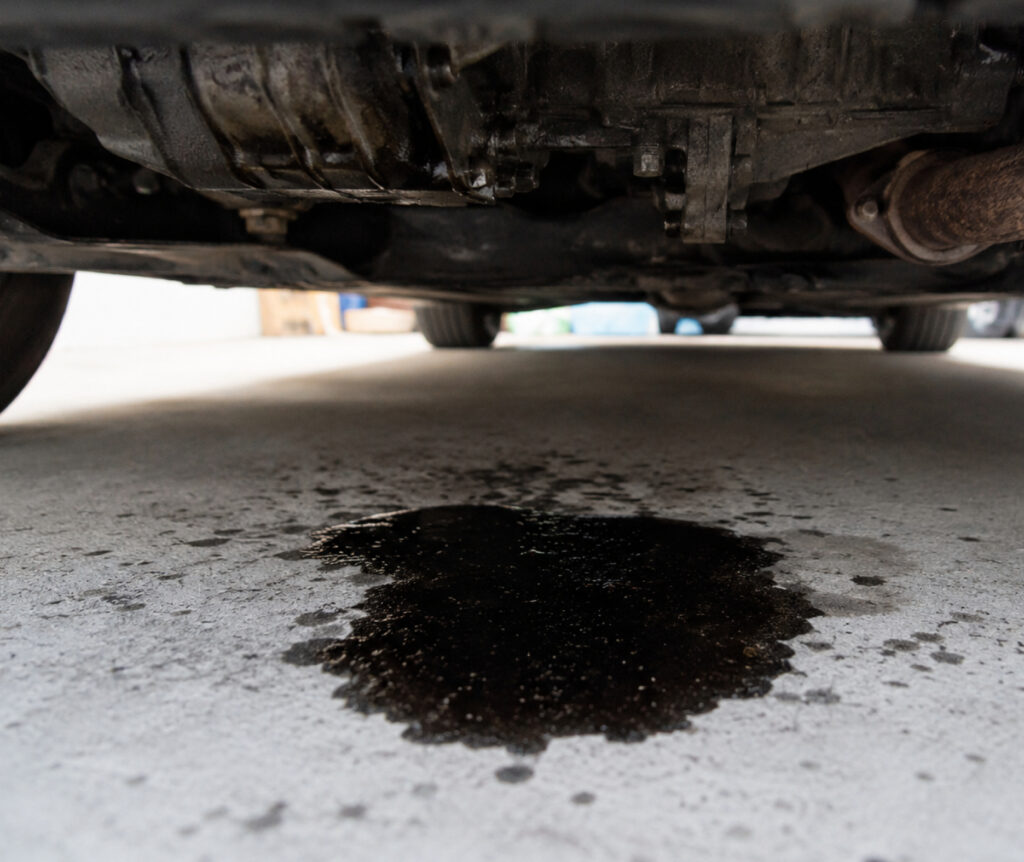 Ground-level view of vehicle undercarriage with oil leak puddle on pavement